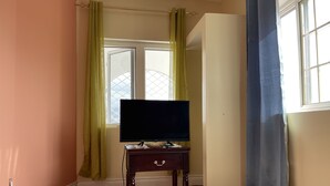 Quarto com televisão sobre mesa de madeira, tapete estampado e janela com cortinas.