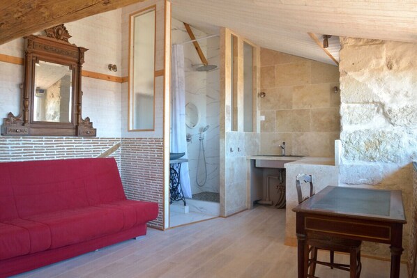 A room with a red sofa, a wooden table, a stone wall, and a bathroom with a shower and sink.