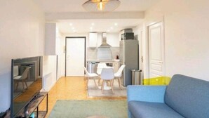 Un saló modern amb un sofà blau, una zona de menjador amb cadires blanques, i una cuina amb armaris blancs i una campana extractora d'acer inoxidable.
