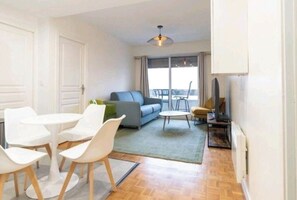 Un salon moderne avec un canapé gris, des chaises blanches, une petite table ronde et un téléviseur posé sur un meuble.