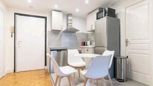 Una cuina compacta amb armaris blancs, una campana extractora d'acer inoxidable, un frigorífic i una petita zona de dinar amb cadires blanques.