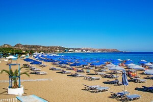 A beach with numerous blue umbrellas and sun loungers, a sandy shore, and a clear view of the sea and distant hills.