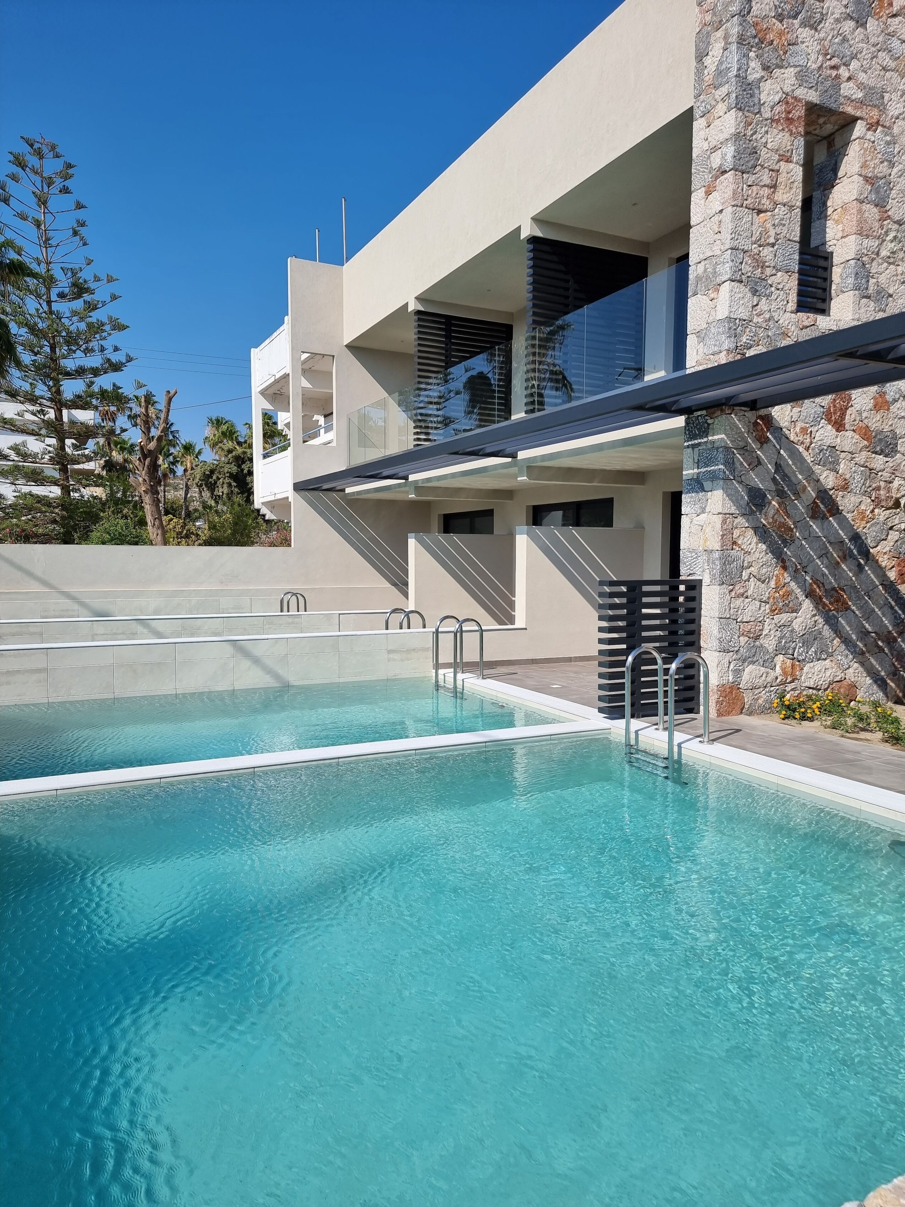 A modern pool area with clear blue water, a stone wall, and a staircase leading to a balcony.