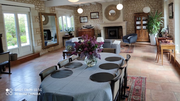 A spacious living room with a fireplace, stone walls, and a dining area with a table set for four. - Chambre vue sur le Jardin (Rivière)