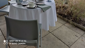 An outdoor dining area with a table set for two, surrounded by greenery and a wooden structure in the background. - Chambre vue sur le Jardin (Rivière)