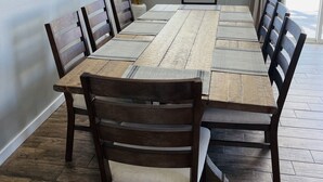 A dining room with a long wooden table and dark wooden chairs, a wall clock, and a window with blinds. - Beautiful Desert Retreat - Salt Water Pool & Spa (Desert Hot Springs)