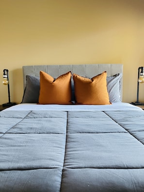 Una cama bien hecha con un cabecero gris y dos almohadas, una naranja y otra gris, frente a una pared amarilla.