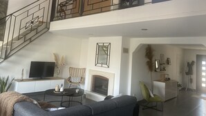 A spacious living area with a high ceiling, a staircase, a fireplace, and a television on a cabinet. - Villa Tout Confort Entre MER ET Etang (Sète)