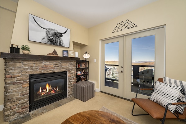 A living room with a stone fireplace, a large framed picture, a bookshelf, and a view through a glass door to the outdoors. - Pool & Pedals Biking Base at Granby Ranch (Granby)