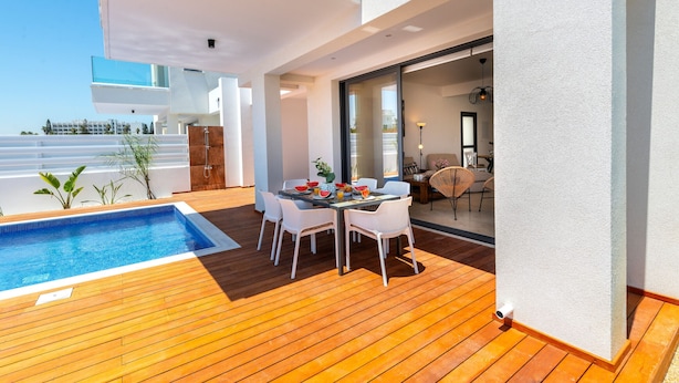 Une terrasse extérieure moderne avec une piscine, une plateforme en bois et un coin repas agrémenté de chaises blanches et d’une table décorée de fruits et d’un vase.