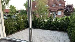 Näkymä lasiovesta sisäpihalle, jossa on kivetty alue. Sisäpihaa ympäröi vehreää kasvillisuutta, ja taustalla näkyy tiilinen rakennus.