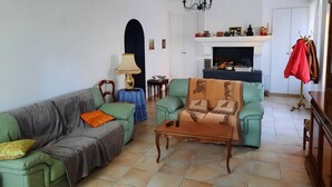 A living room with a sofa, armchair, and coffee table. There is a fireplace with a mantel, a lamp, and a coat rack. - Villa near beach, golf course, verdon gorges (Trans-en-Provence)