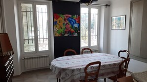 A dining room with a round table covered in a floral tablecloth, surrounded by wooden chairs. There are two windows with white frames, a radiator below the windows, and a colorful abstract painting on the wall. - Villa near beach, golf course, verdon gorges (Trans-en-Provence)
