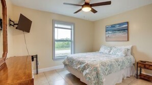 Un dormitorio con cama, ventilador de techo, ventana y un cuadro en la pared.