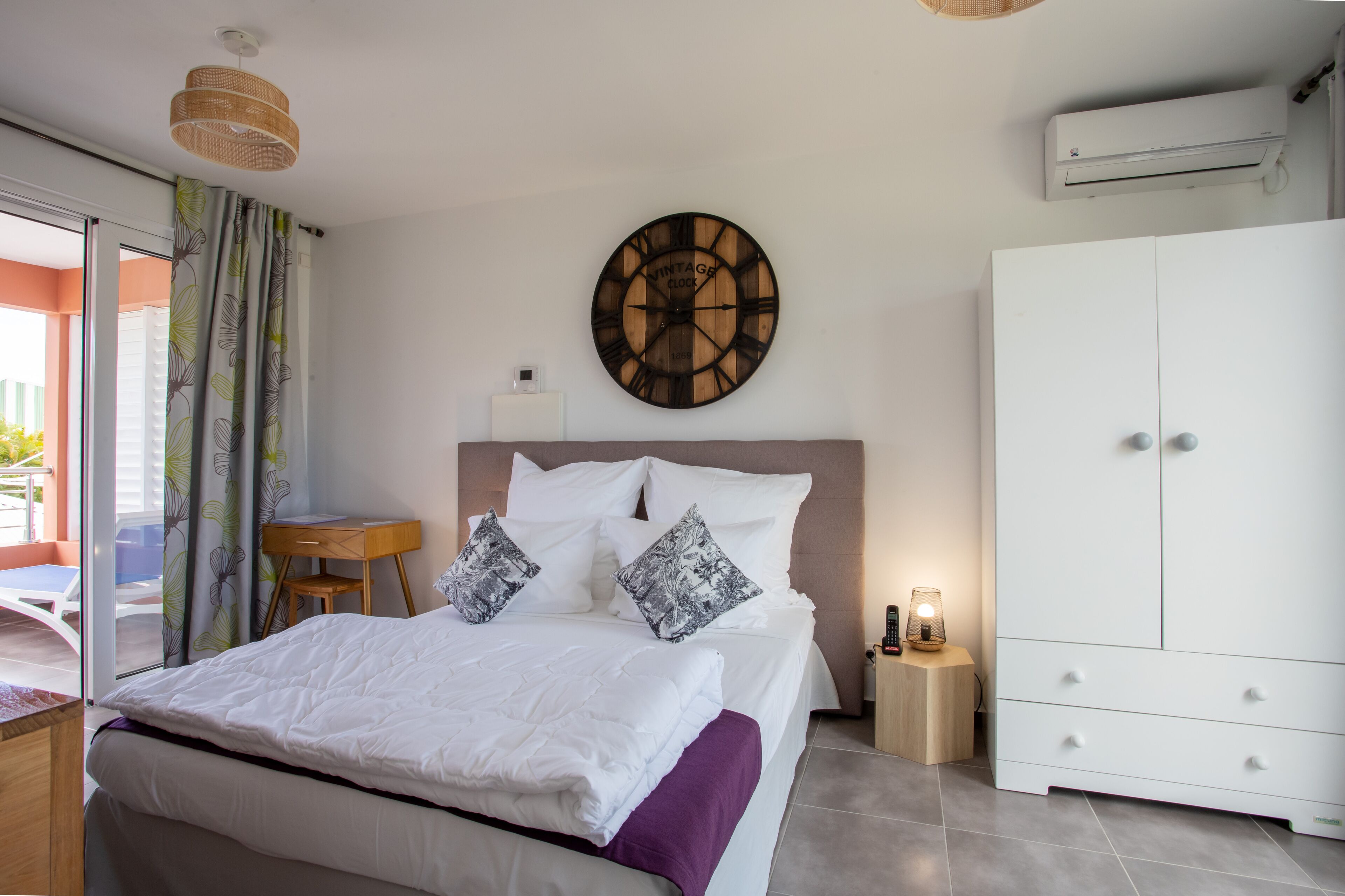 A bedroom with a bed, a large white wardrobe, a wall clock, a bedside table with a lamp, and a window with curtains.