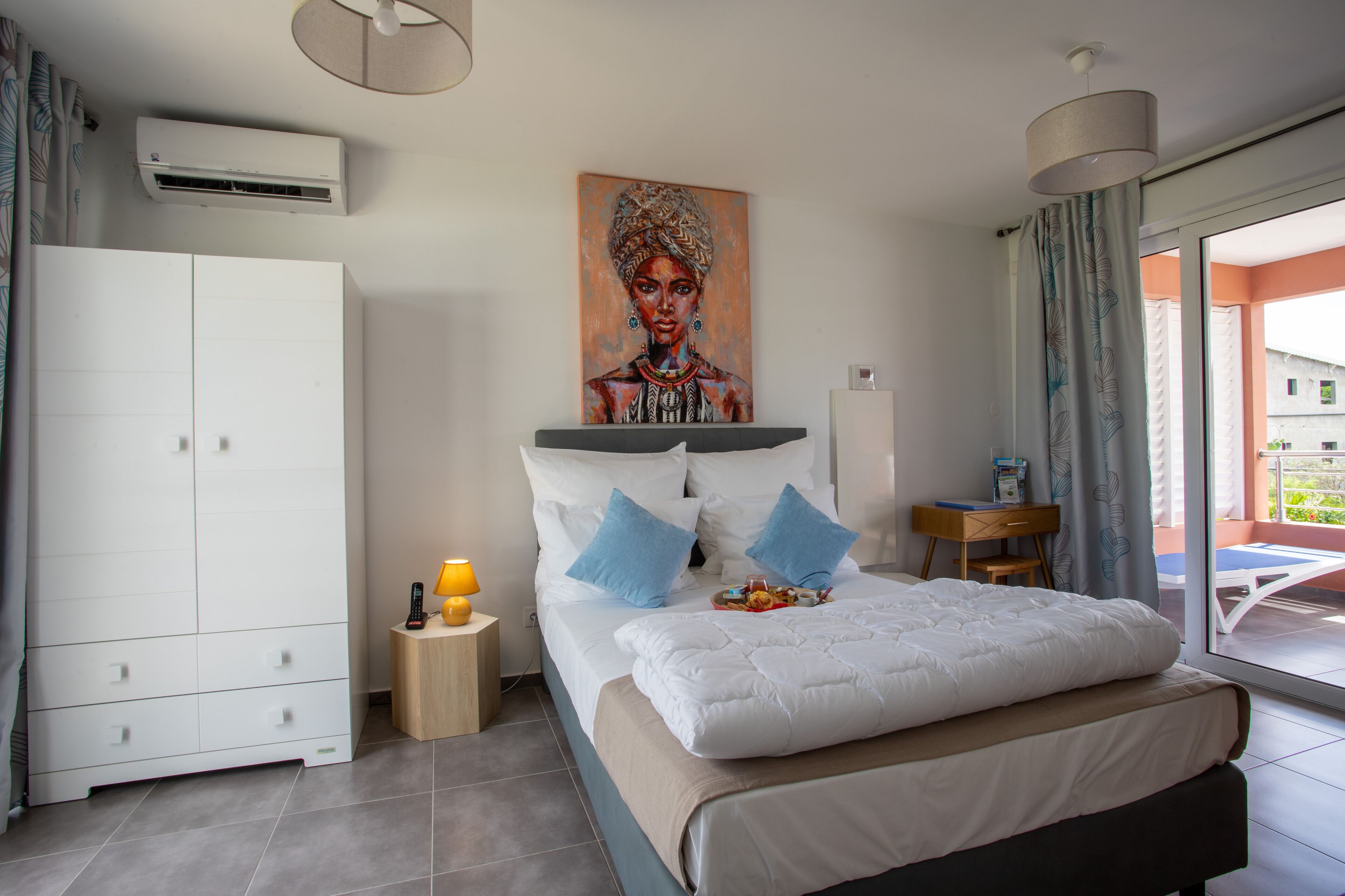 A bedroom with a bed, a wardrobe, a painting, a desk, and a view of the outside.