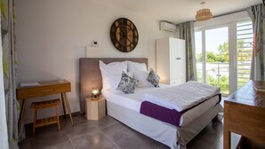 A bedroom with a bed, bedside table, desk, and a wall clock. - Hôtel Guadeloupe Palm Suites (Saint-François)
