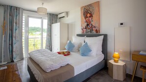 A modern hotel room with a large bed, a painting on the wall, and a view of greenery outside. - Hôtel Guadeloupe Palm Suites (Saint-François)