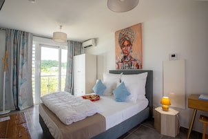 A modern hotel room with a large bed, a painting on the wall, and a view of greenery outside.