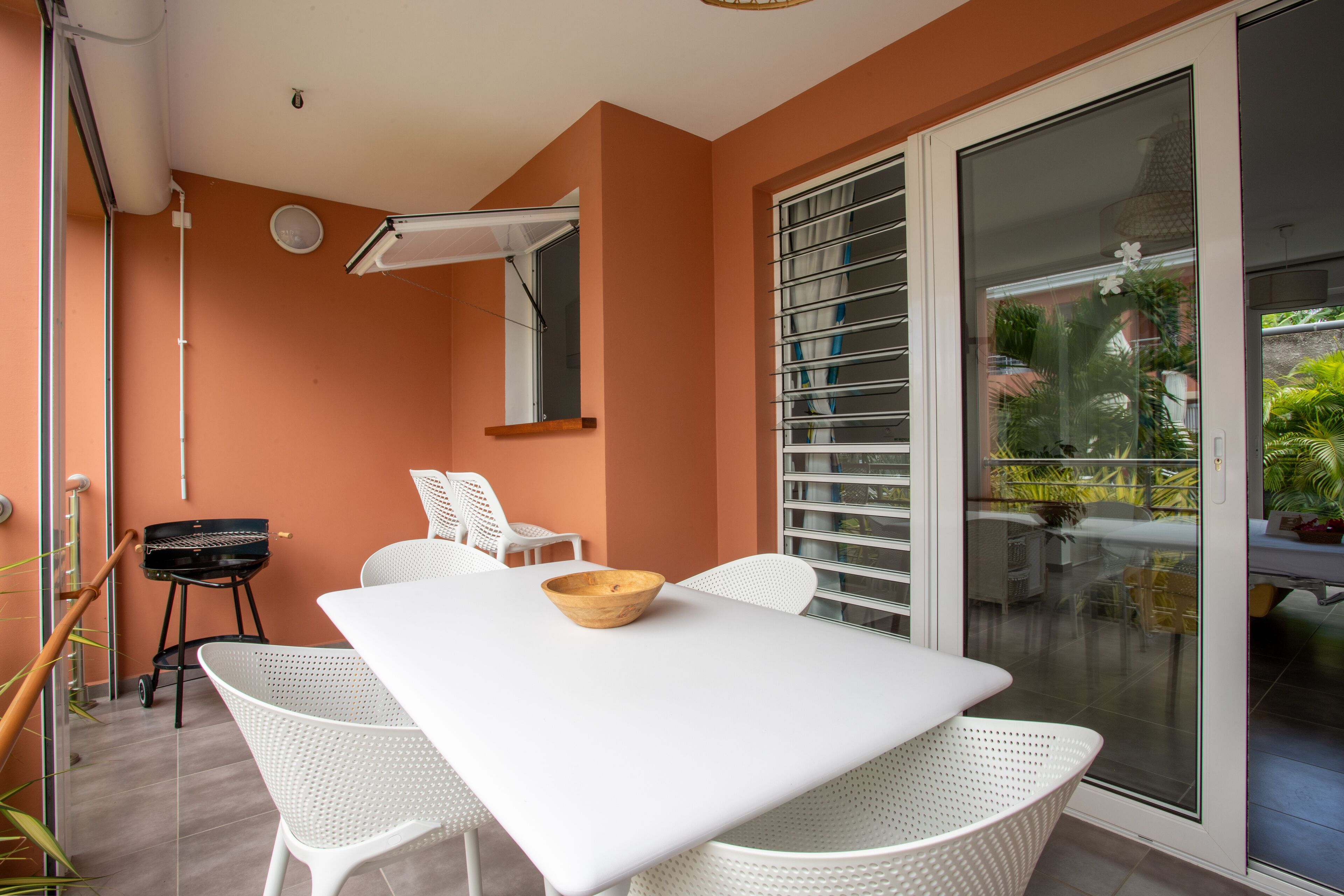 A dining area with a white table and chairs, a wooden bowl, and a sliding glass door leading to a balcony with plants.