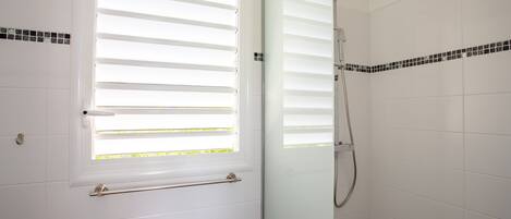 A bathroom with white tiled walls, a glass shower enclosure, and a window with horizontal blinds.