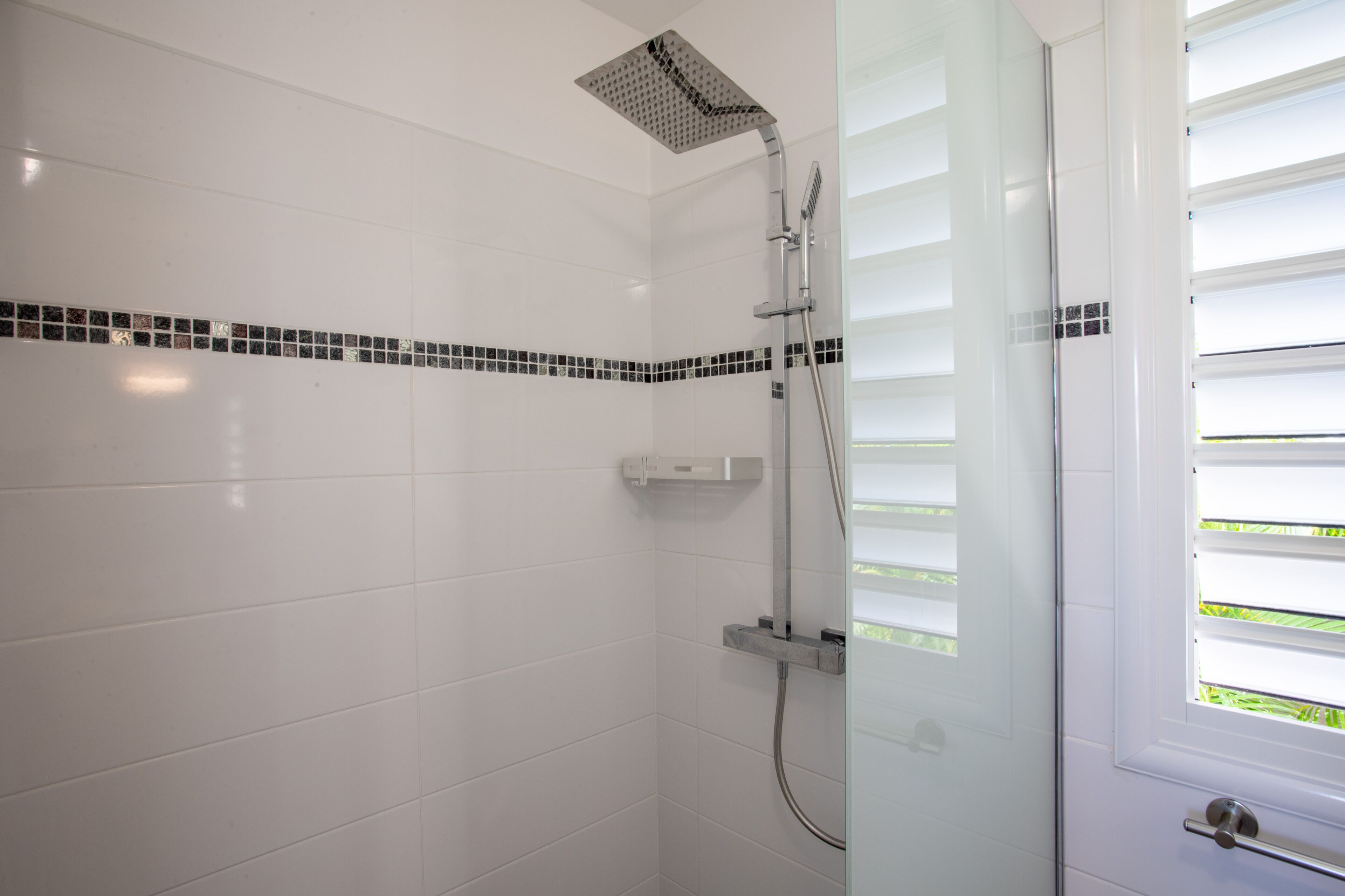 A modern bathroom with white tiled walls, a shower with a square head, and a window with blinds.