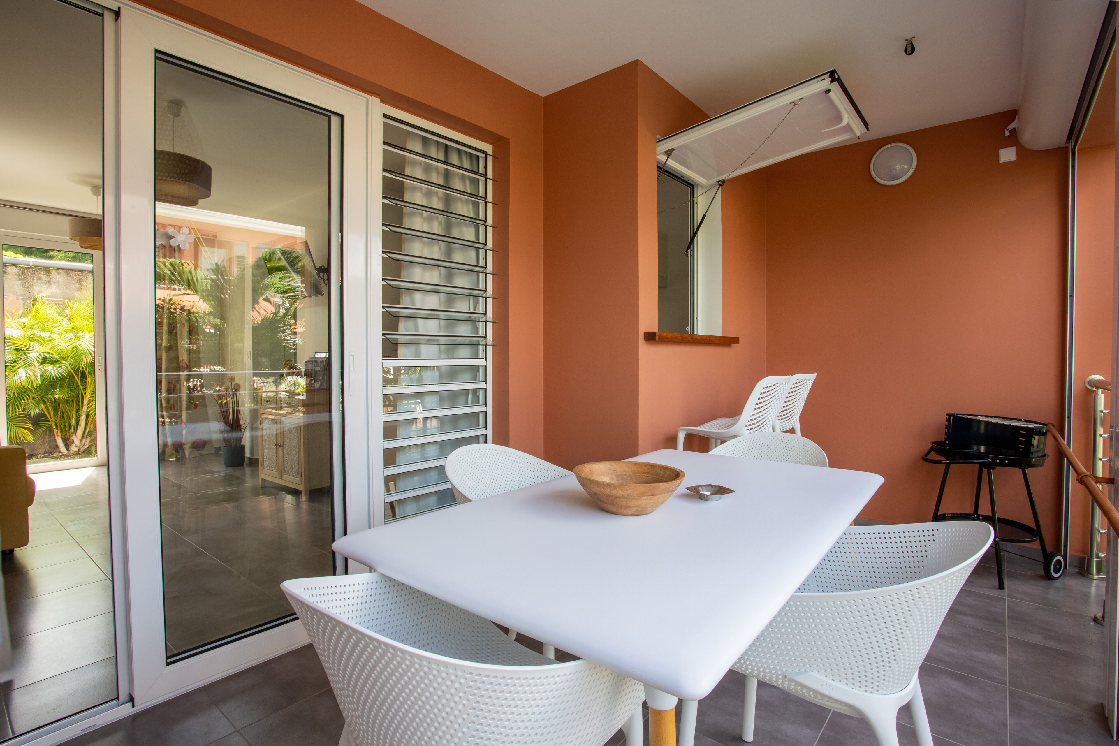 Uma sala de jantar moderna com uma mesa e cadeiras brancas, uma tigela de madeira e uma porta de vidro deslizante que dá para um pátio externo.