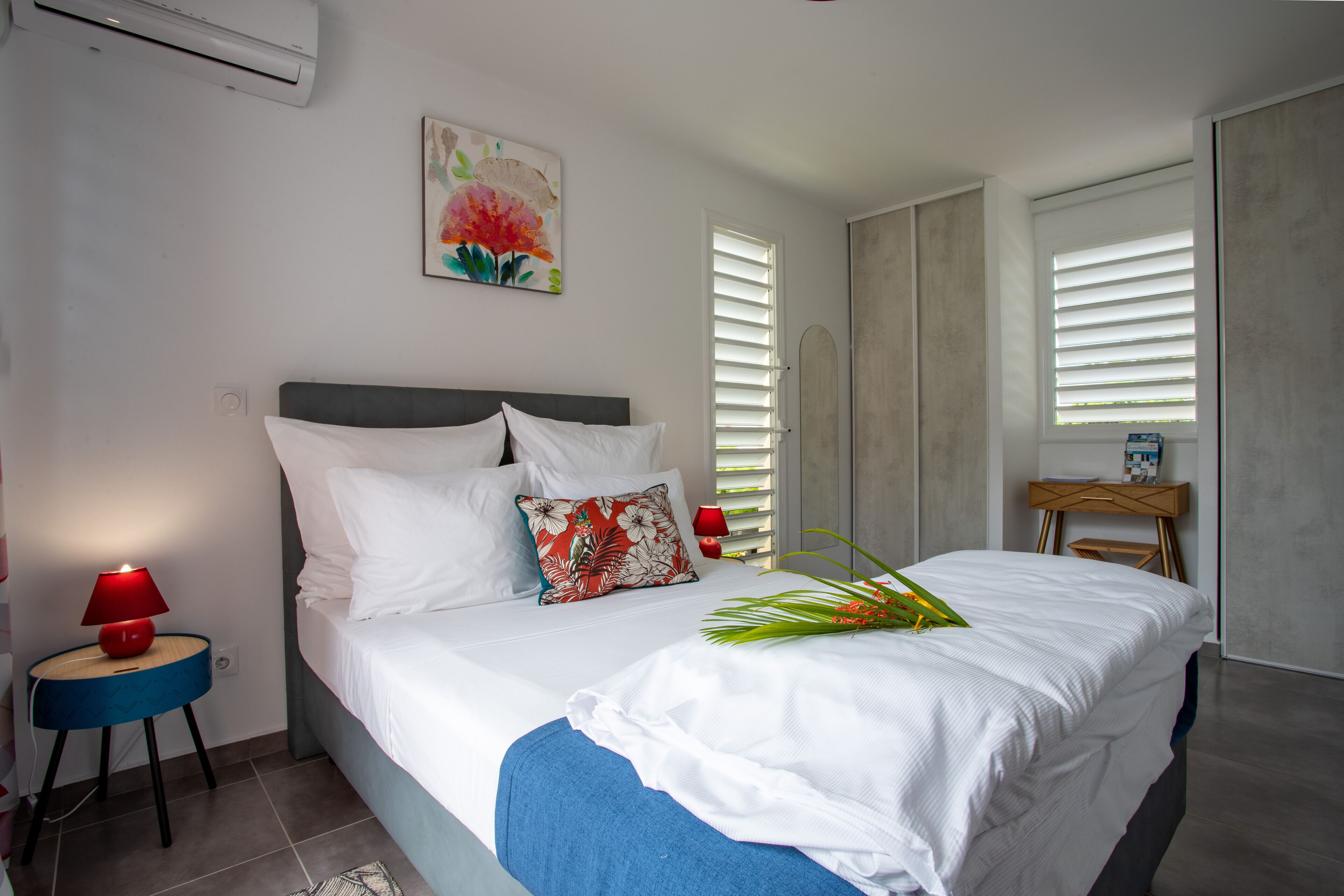 Un dormitorio con cama, mesita de noche, lámpara, un cuadro y ventana con persianas.