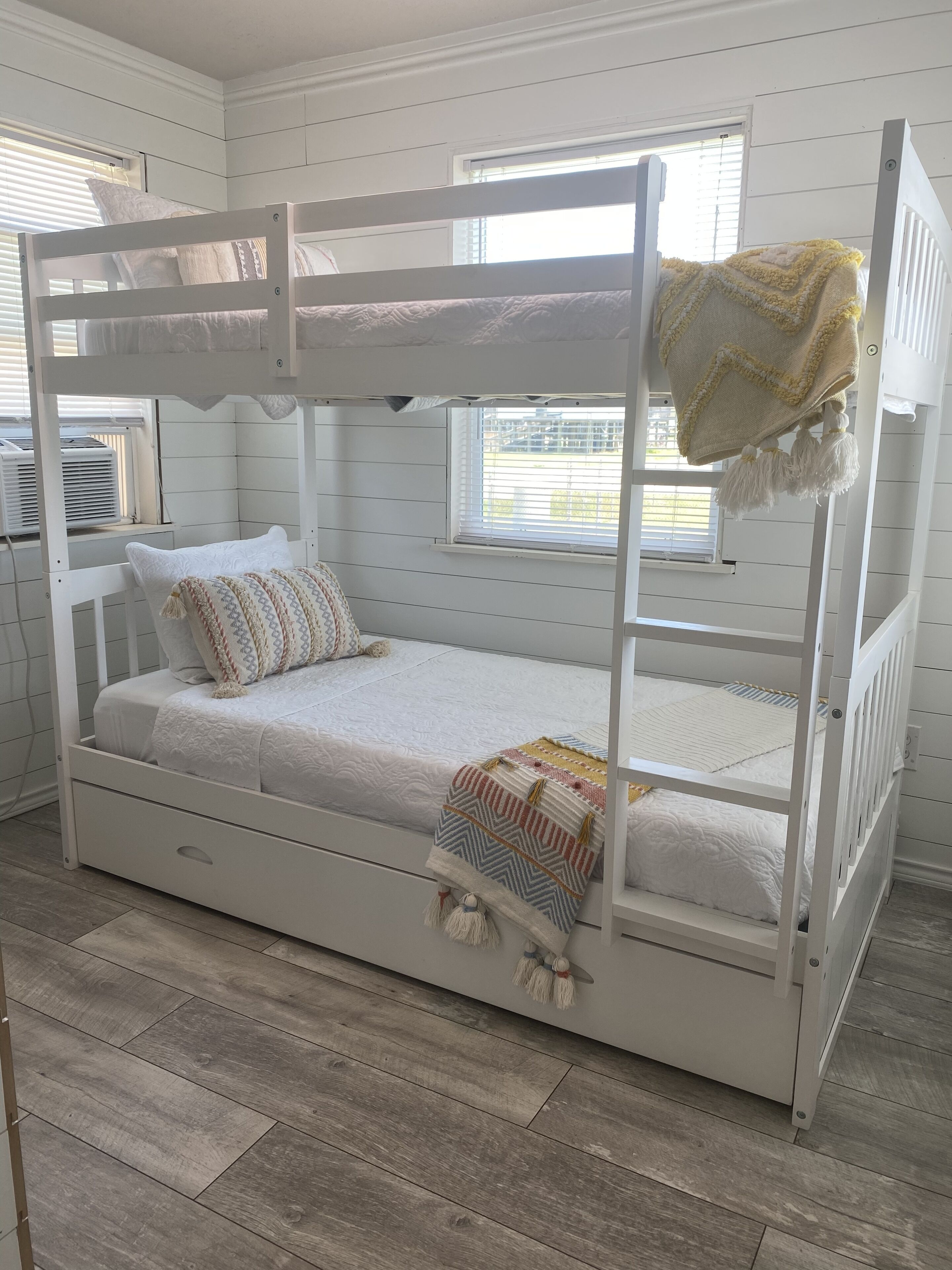 A bunk bed with a white frame, a lower bed with a patterned blanket, and a window with blinds.