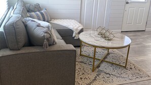 Uma sala de estar com sofá seccional cinza, mesa de centro redonda com pés dourados, tapete estampado e uma grande obra de arte abstrata dourada na parede.