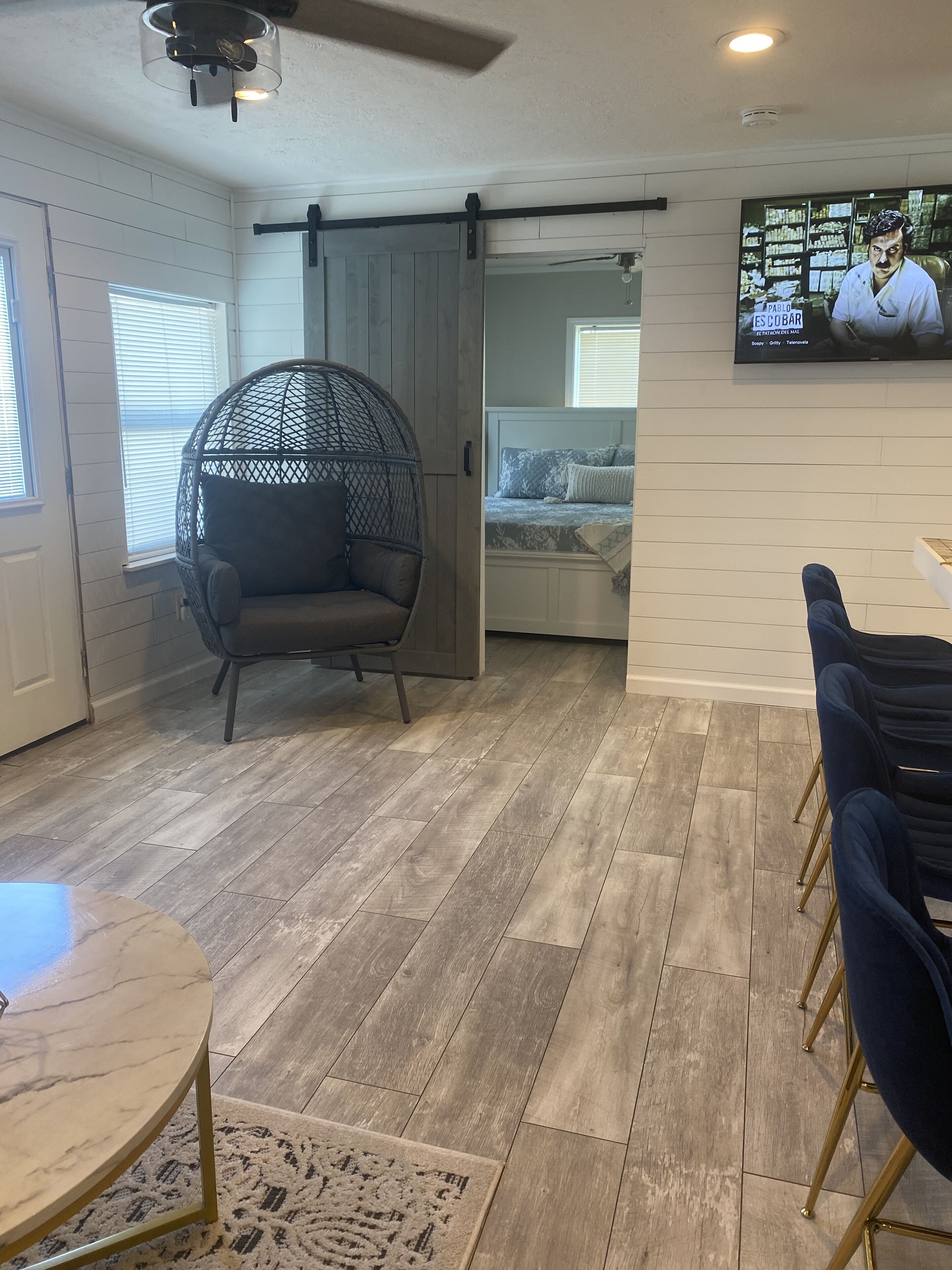 A room with a wooden floor, a round table, a hanging chair, and a bed with a TV in the background.