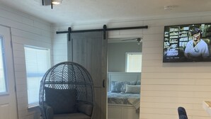 Quarto com piso de madeira, uma mesa redonda, uma cadeira suspensa e uma cama com televisão na parede.
