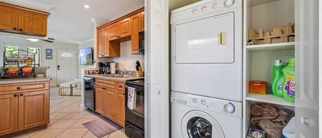 Une buanderie équipée d’une machine à laver et d’un sèche-linge superposés, d’une étagère contenant des produits de nettoyage, et un coin cuisine avec des meubles en bois et un four à micro-ondes.