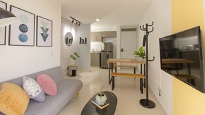 Una sala moderna con un sofá gris, una mesa de centro y un televisor. Se observa una zona de comedor con una mesa y sillas de madera. Se ve la cocina al fondo, con un refrigerador y un lavamanos.