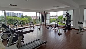 Un gimnasio amplio con diversos equipos de ejercicio, ventanas grandes y un balcón con vistas a zonas verdes y edificios.