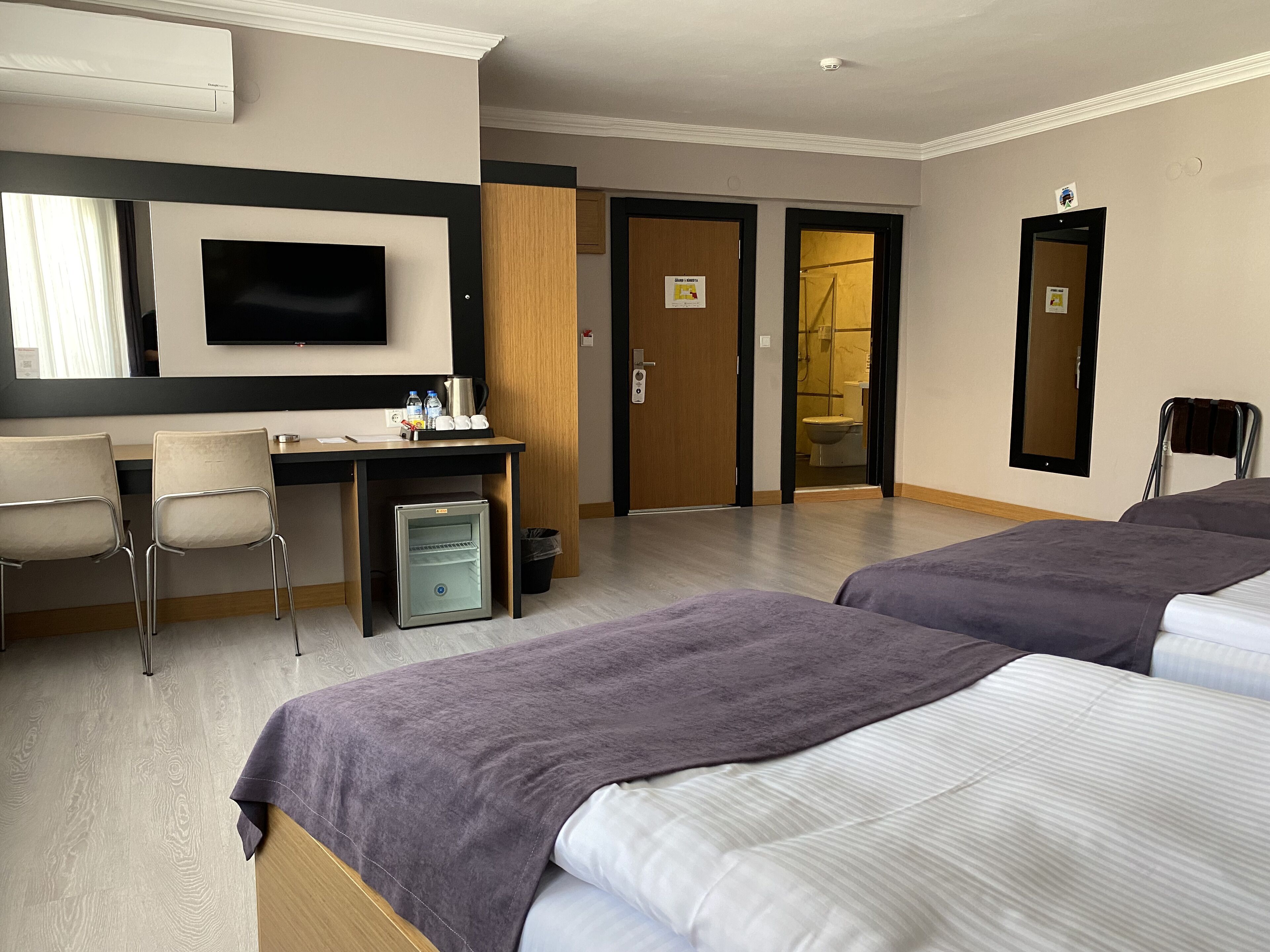 A hotel room with two beds, a desk with chairs, a television, and a small refrigerator.