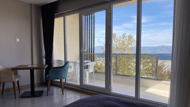 Een hotelkamer met een grote schuifglazen deur die uitkomt op een balkon met uitzicht op zee en bergen. In de kamer staat een bed, een stoel en een klein tafeltje.