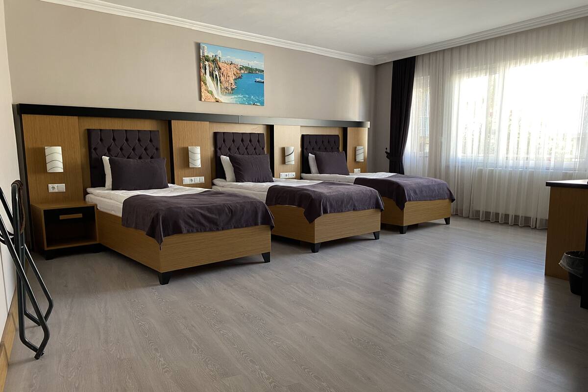A hotel room with four beds, a large painting of a coastal scene, and a large window with sheer curtains.