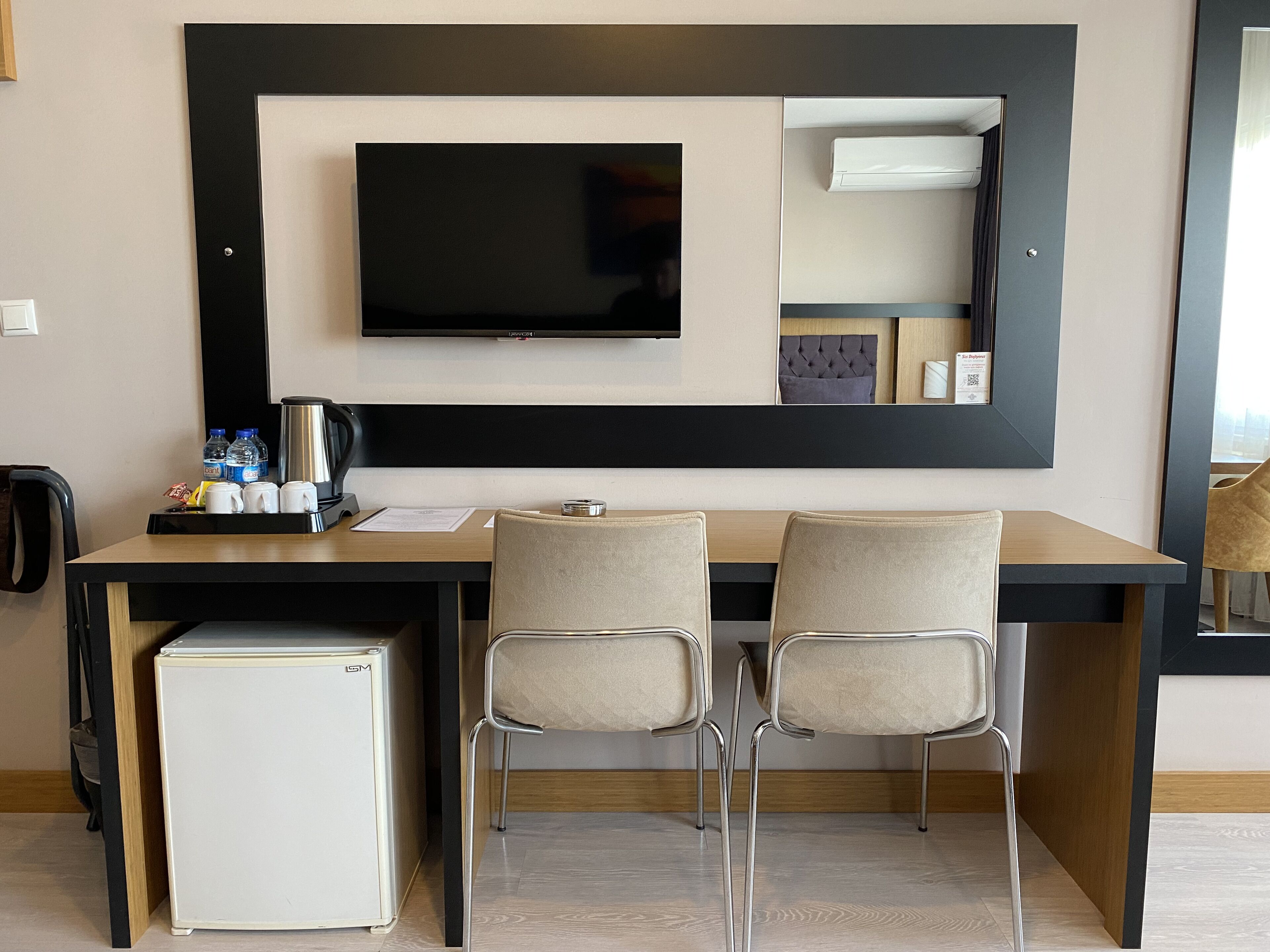 A hotel room with a desk, two chairs, a small fridge, a television, and a mirror.