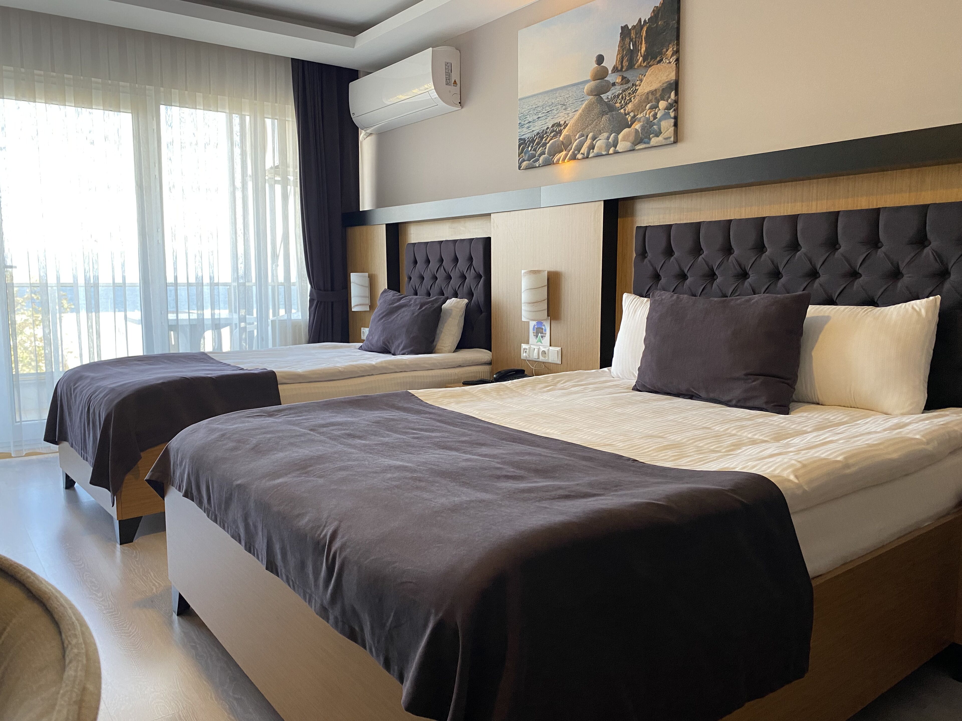 A hotel room with two beds, a large window with curtains, a wall-mounted air conditioner, a painting of a coastal scene, and a wooden headboard.