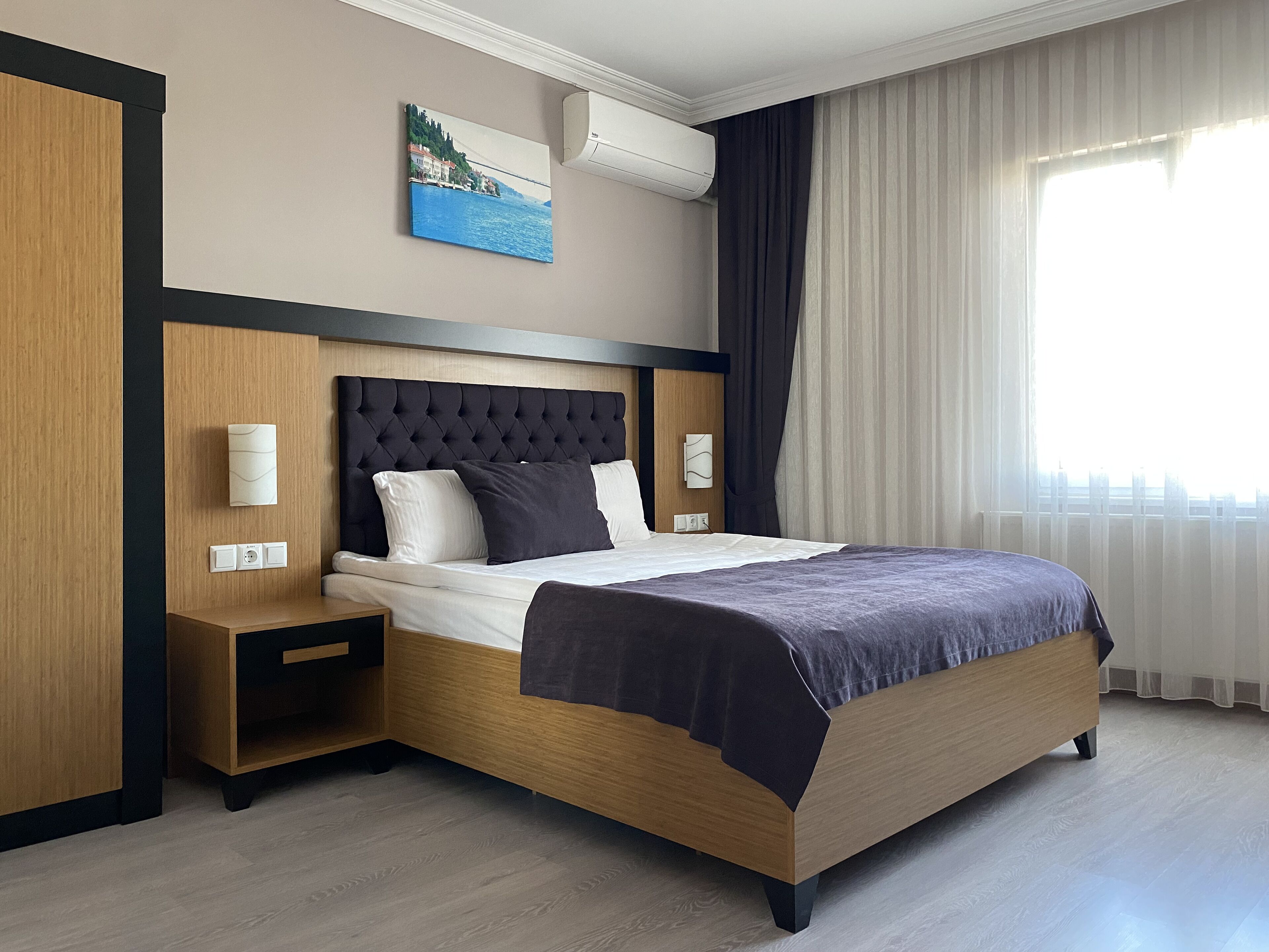 Ett modernt hotellrum med en sänggavel i trä, en stor säng med en mörk sänggavel, ett nattduksbord med en lampa och ett fönster med gardiner.