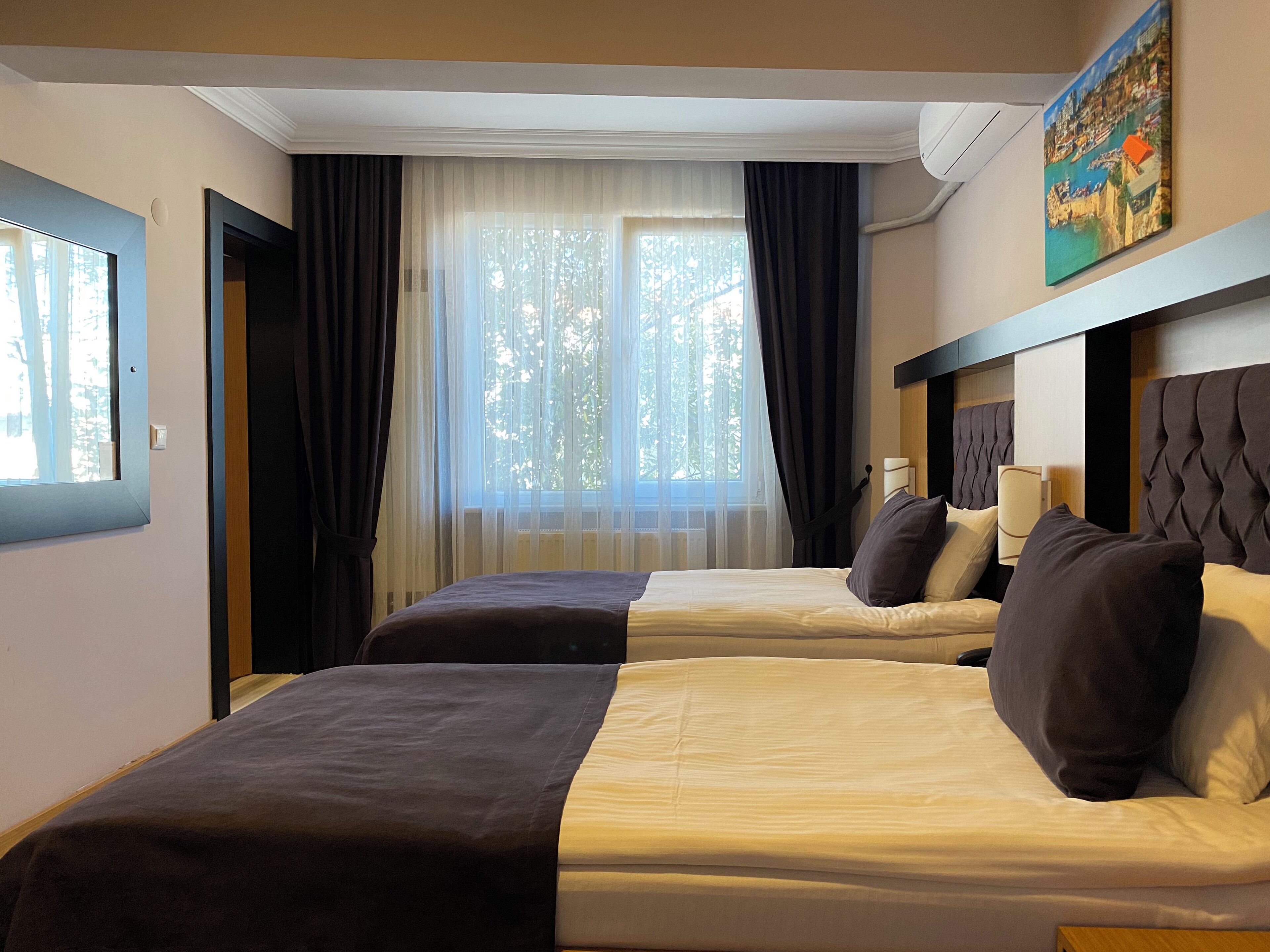 Ein Hotelzimmer mit zwei Betten, einem großen Fenster mit leichtem Vorhang und einem Blick auf eine Küstenstadt.