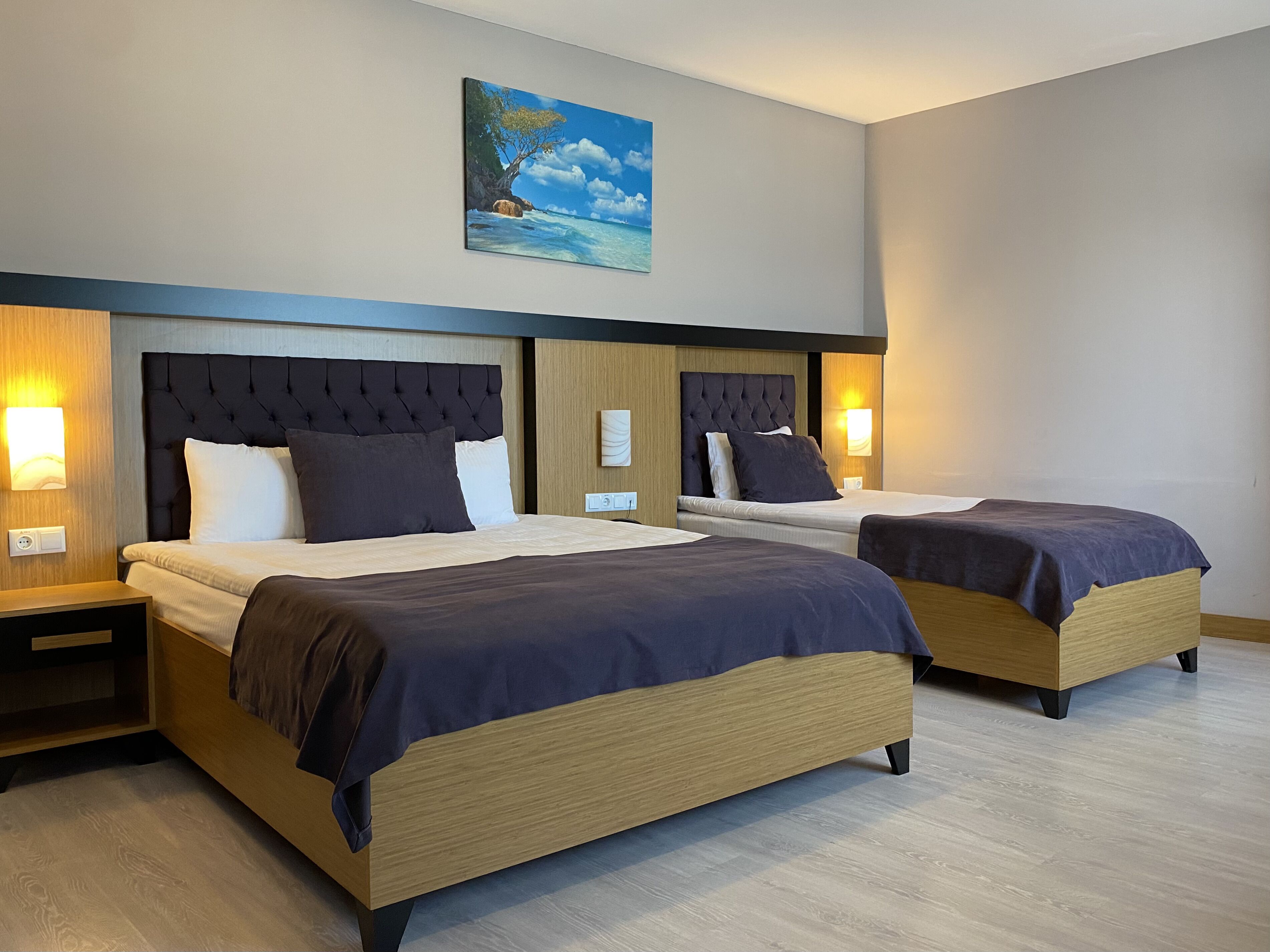Habitación de hotel con dos camas, cada una con cabecero de madera y cubrecama azul oscuro. Hay mesitas de noche con lámparas y un cuadro con una escena de playa en la pared.