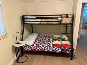 Un lit superposé avec une courtepointe à motifs, une petite table ronde avec une lampe et une fenêtre avec des stores.