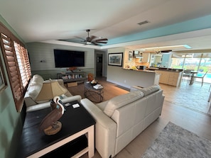 A modern living room with a sofa, coffee table, and TV. A kitchen with a bar and a dining area is visible in the background.