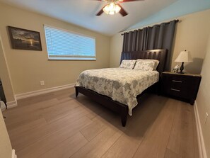 A bedroom with a bed, a nightstand, a ceiling fan, a window with blinds, and a wall-mounted picture.