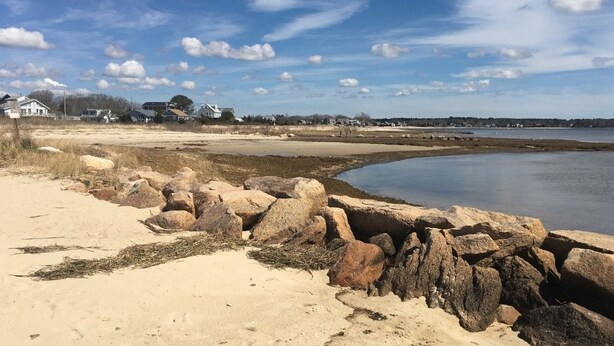 Un paysage côtier avec une plage de sable, de gros rochers et une étendue d’eau calme.