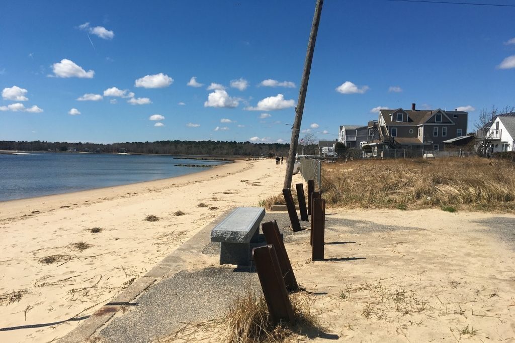 Una playa con un banco de hormigón, postes de madera y casas al fondo.