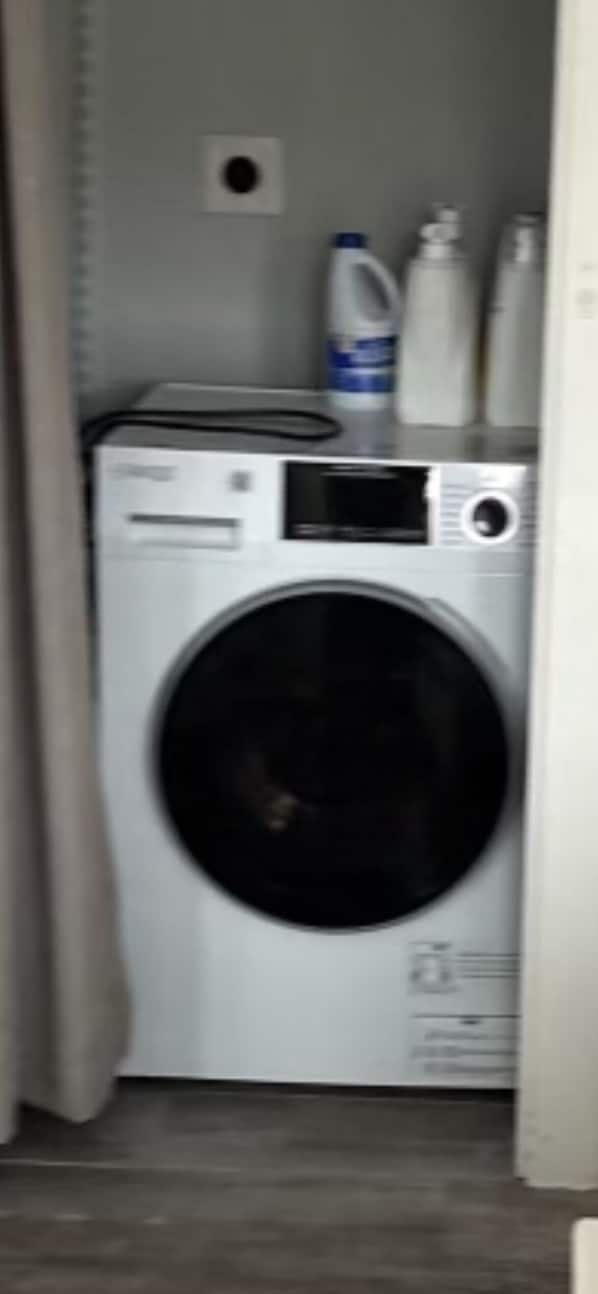 Une petite buanderie moderne et fonctionnelle, équipée d’un lave-linge à chargement frontal, d’un sèche-linge et d’une petite étagère où sont rangés les produits de nettoyage.