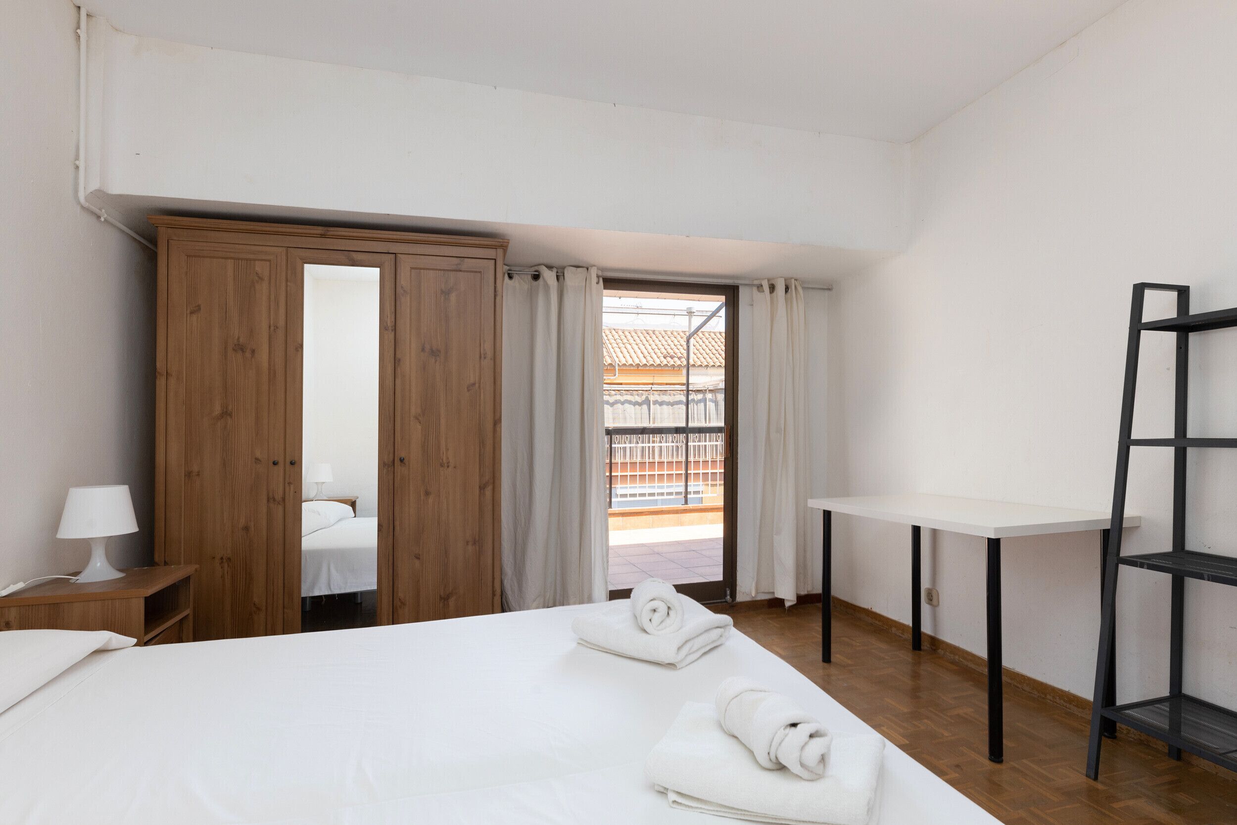 Een slaapkamer met een bed, een houten garderobe, een bureau, een stoel en uitzicht op een balkon.
